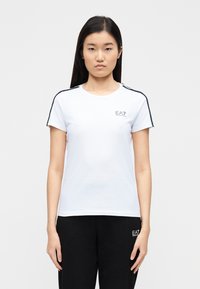 Balta īsroku T-krekls ar apaļu kakla izgriezumu, uz pleciem ar melniem un sarkaniem svītru akcentiem un nelielu EA7 logotipu uz krūtīm.