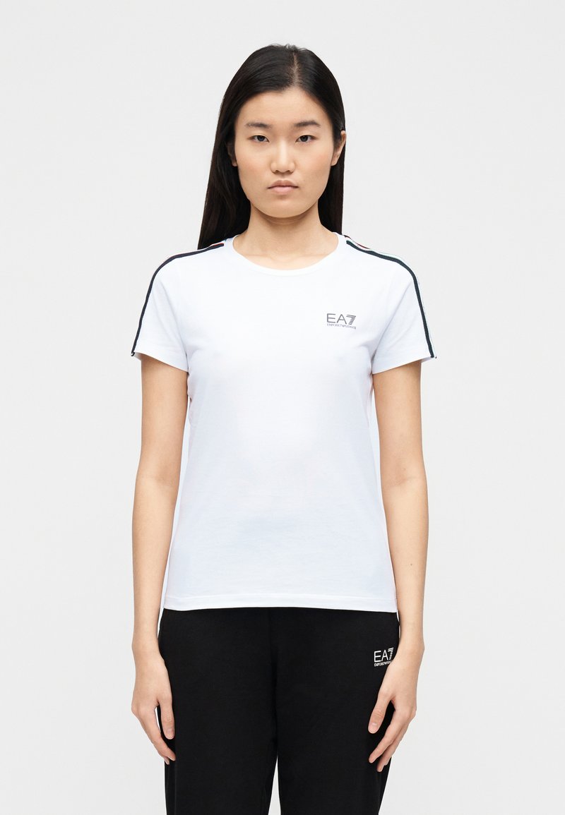 Balta īsroku T-krekls ar apaļu kakla izgriezumu, uz pleciem ar melniem un sarkaniem svītru akcentiem un nelielu EA7 logotipu uz krūtīm.