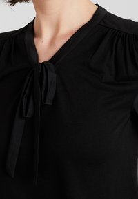 Svart topp med v-ringning och knytning vid kragen. Mjukt material med lätt rynkning vid axlarna. Enkelt, minimalistiskt design.