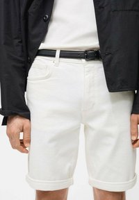 Pantaloni corti bianchi in cotone con risvolti, dotati di tasche frontali e vestibilità dritta, indossati con una giacca nera e una cintura nera.