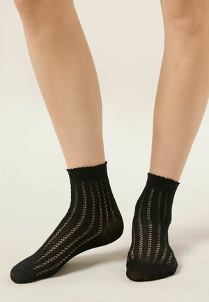 Jambes portant des chaussettes courtes noires avec un motif fin en maille ouverte, debout sur un fond clair uni.