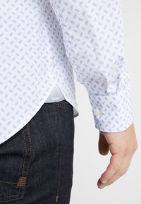 Vit skjorta med små mönster av blå blad, tillverkad av bomull, med rundad nederkant. Kombineras med mörka denimjeans som visar en logotag på nederkanten.