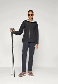 Μαύρο ελαφρύ μπουφάν με φερμουάρ, συνδυασμένο με ναυτικές cargo παντελόνες και καφέ παπούτσια πεζοπορίας. Το μοντέλο κρατά μαύρα trekking poles και φορά γυαλιά ηλίου.