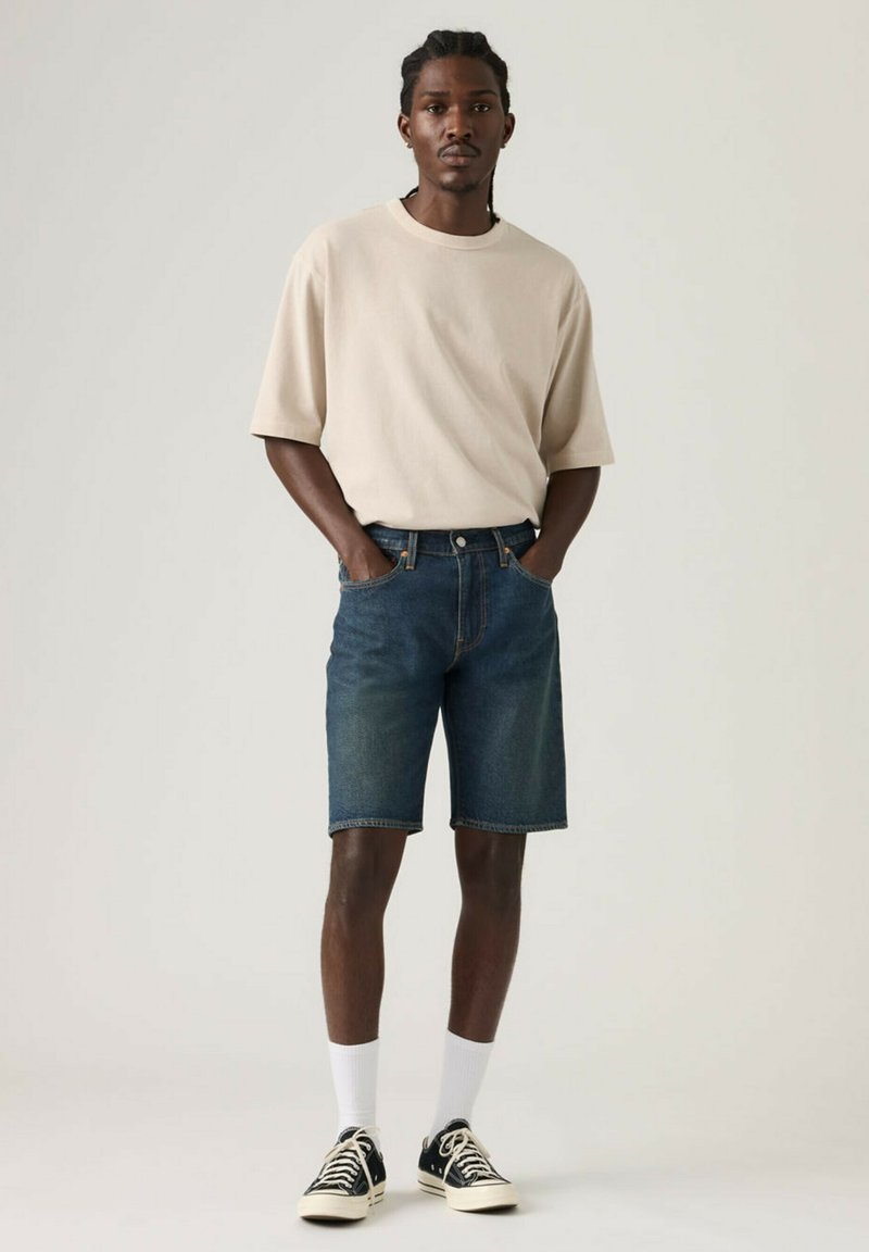 Jeune homme debout les mains dans les poches, portant un t-shirt beige, un short en jean foncé, des chaussettes blanches et des baskets noires sur un fond uni.