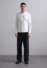 Hvid langærmet skjorte med lille sort logo på brystet, parret med sorte løse bukser og sorte sneakers, sat mod en neutral baggrund.