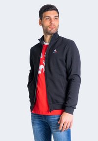 Chaqueta negra con cremallera y alto cuello, puños acanalados y logo en el pecho, llevada sobre una camiseta roja y jeans azules.