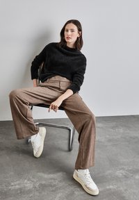 Pull noir en peluche avec un large col, pantalon à carreaux marron taille haute, et baskets blanches avec des accents gris, assis sur une chaise en métal.