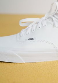 White canvas sneaker with a low-top design, rounded toe, and flat cotton laces. Features a small Vans logo tag on the side. Textured rubber sole.