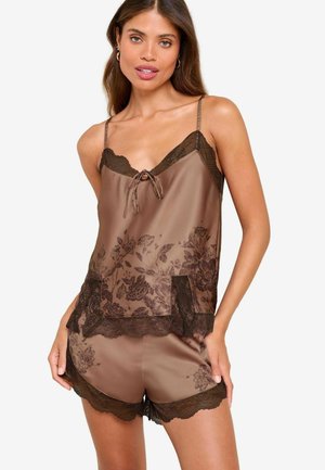 Nő barna selyem camisole és rövidnadrág pizsamában, virágos és csipkés díszítéssel, fehér háttér előtt pózol.