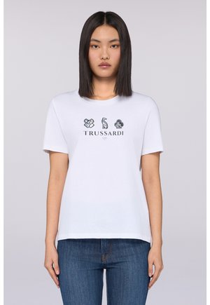 Camisa de algodón blanca con logotipos impresos en negro en la parte frontal, cuello redondo y mangas cortas. Combinada con jeans de mezclilla azul.