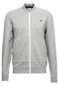 Šedá mikina na zip, vyrobená z měkké tkaniny, má žebrovaný límec, manžety a spodní lem. Na hrudi je malé zelené logo.