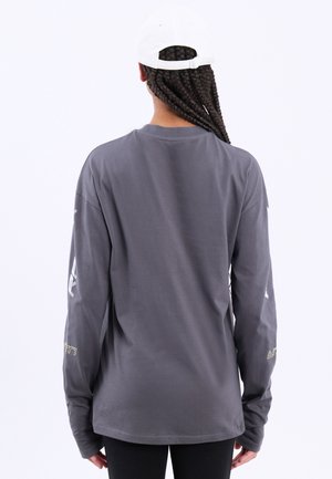 Personne aux longs cheveux tressés portant une casquette blanche et un t-shirt gris à manches longues, vue de dos sur un fond blanc uni.