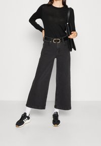 Haut noir à manches longues, jeans en denim noir à jambes larges, boucle de ceinture en or et baskets noires avec des accents blancs. Tenant une pochette noire.