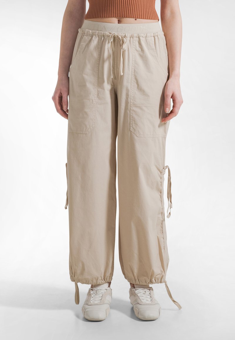 Beige cargobukser med justerbare snøre ved manchetterne, elastisk talje og forlommer med lapper foran. Fremstillet af let, struktureret stof.