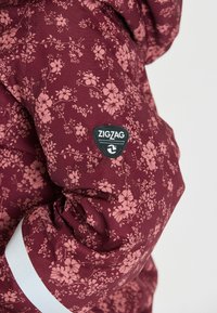 Bordeaux winterjack met een bloemenpatroon in het roze, voorzien van een zwart Zigzag-logo patch en een witte accentstreep op de mouw.