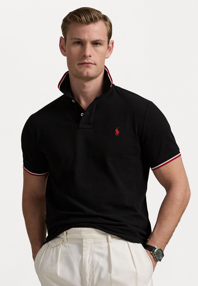 Homem a usar uma camiseta polo preta com detalhes em vermelho e branco, calças brancas e um relógio de pulso preto, de pé contra um fundo branco simples.