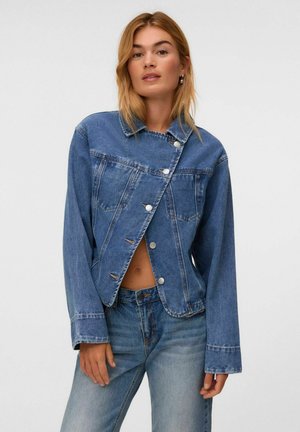 Femme portant une veste en jean bleue asymétrique boutonnée et un jean taille haute assorti, posant avec une expression neutre sur fond blanc.