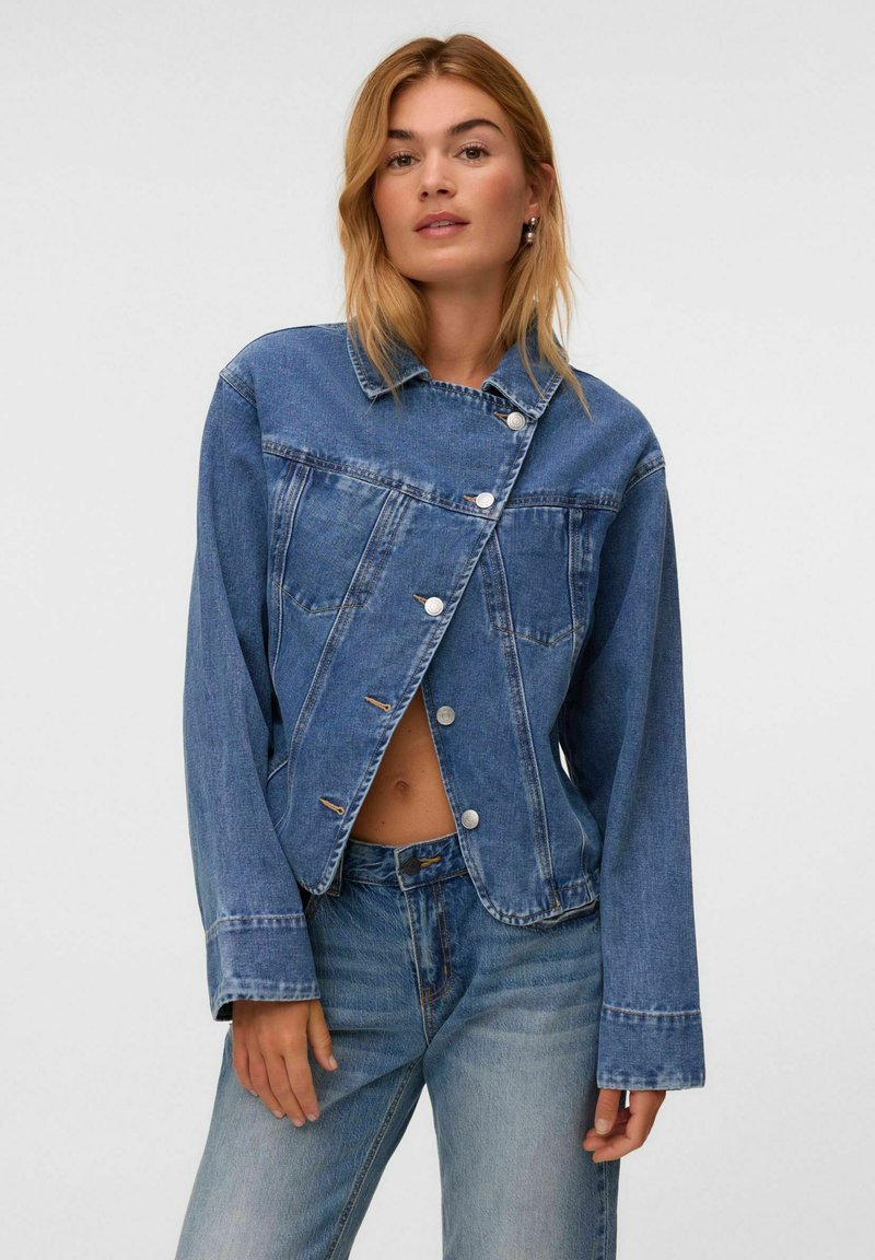 Femme portant une veste en jean bleue asymétrique boutonnée et un jean taille haute assorti, posant avec une expression neutre sur fond blanc.