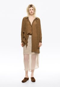 Camisa de botones marrón y texturizada sobre una falda beige con flecos y una capa translúcida; combinada con zapatos marrones de tipo slip-on.