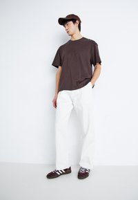Bruine katoenen t-shirt, relaxte pasvorm, korte mouwen. Gecombineerd met witte wijd-uitlopende broek en donkerpaarse sneakers met witte strepen.