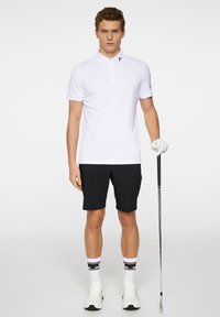 Valkoinen polo-paita lyhyillä hihoilla, mustilla yksityiskohdilla ja kaulusnapilla. Yhdistetty mustiin shortseihin, valkoisiin kenkäavain ja golfmaila kädessä.