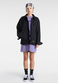 Czarna, oversize kurtka z dwiema przednimi kieszeniami, noszona na lila sukience. Model nosi wzorzystą czapkę, czarne skarpety i buty Vans.