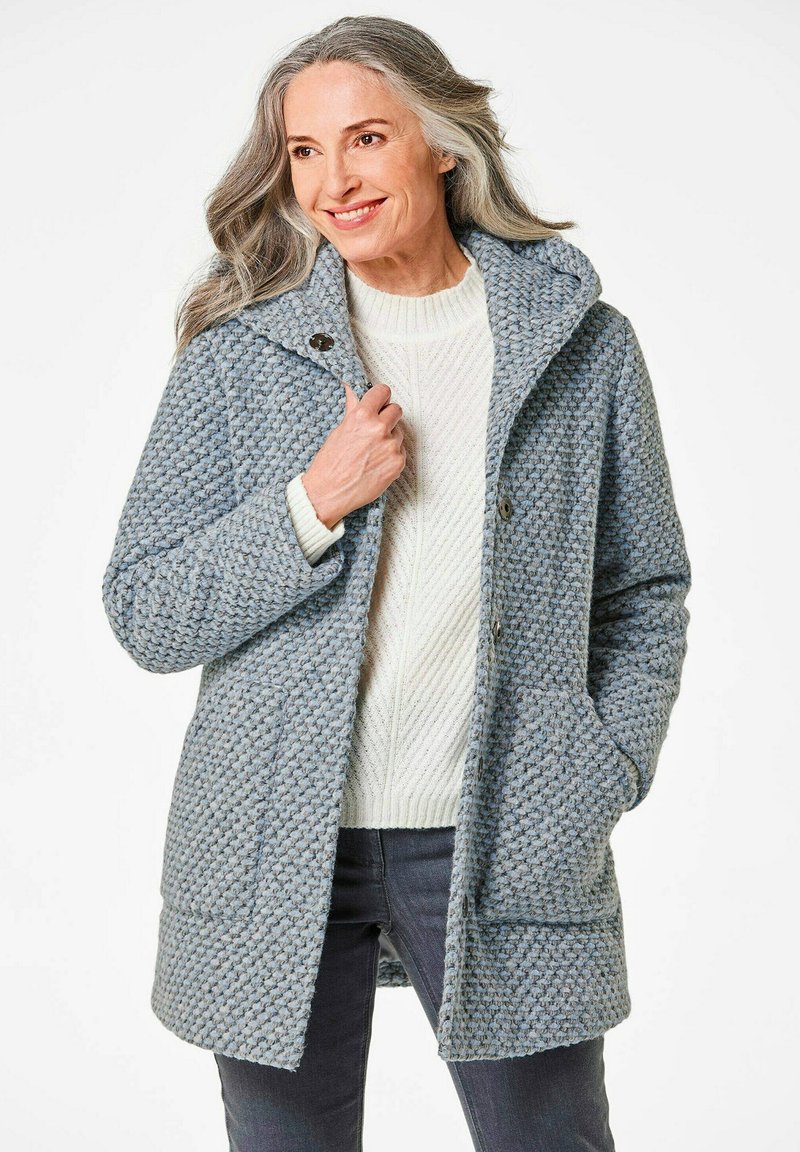 Himmelblauer, strukturierter Mantel mit Kapuze und zwei vorderen Taschen, mit Knopfverschluss und gestricktem weißen Pullover darunter.