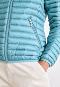Gaiši zils pūkains jaka ar quilted tekstūru, sudraba rāvējslēdzējiem un logotipa lentu apakšmalā, apvienota ar gaiši bežām biksēm.