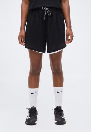 Osoba ubrana w czarne sportowe spodenki z białym obszyciem, białe skarpety Nike do kostki oraz czarne buty sportowe Nike, stojąca na białym tle.