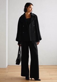 Manteau noir à double boutonnage avec des revers larges, associé à un top noir et un pantalon large. Les accessoires comprennent un sac noir et des talons aiguille.
