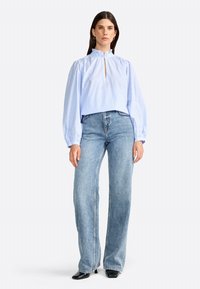 Blouse rayée bleu clair coupée avec un col haut, des manches bouffantes et une fermeture éclair à l'avant. Associée à un jean en denim clair à jambe large et des chaussures noires.