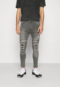 Jeans de denim gris deslavado con detalles desgastados, que presentan grandes secciones rasgadas y un ajuste ceñido, combinados con zapatillas negras y blancas.