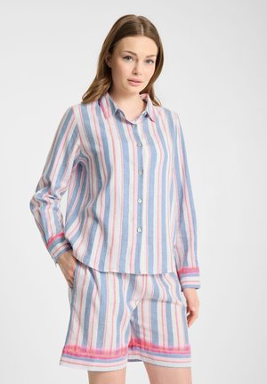 Kobieta ubrana w swobodną koszulę w paski i dopasowane szorty w kolorach niebieskim, różowym i białym, stojąca z jedną ręką w kieszeni.