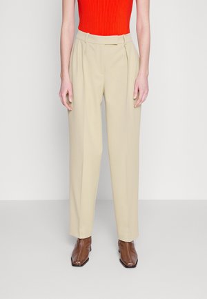 Kobieta ubrana w beżowe, dopasowane spodnie z wysokim stanem, jasny pomarańczowy prążkowany top bez rękawów oraz brązowe botki z kwadratowym noskiem, stojąca na jednolitym tle.