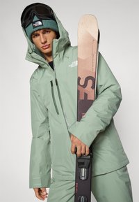 Chaqueta impermeable verde claro con cierre delantero. La persona sostiene un esquí con una parte superior estampada y las fijaciones visibles. Lleva puesta una gorra de lana.