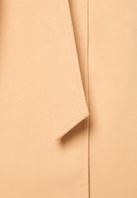 Beżowy materiał, gładka tekstura, z detalem klapki, prosty krój, z czystymi brzegami i minimalnymi akcentami szewnymi.