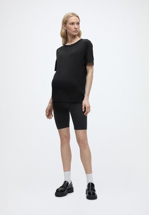 Femme debout portant un t-shirt noir oversize, un short de cycliste noir, des chaussettes blanches et des mocassins noirs épaisses, regardant sur le côté.