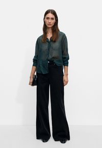 Przezroczysta, ciemnoniebieska koszulabutton-up z długimi rękawami, zestawiona z czarnymi spodniami o wysokim stanie i szerokich nogawkach oraz czarnymi, spiczastymi butami, trzymająca małą torebkę.