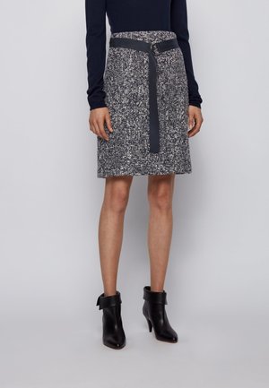 Frau trägt einen knielangen strukturierten Rock mit einem schwarzen Gürtel, ein schwarzes Langarmshirt und schwarze Leder-Stiefeletten mit Absatz.