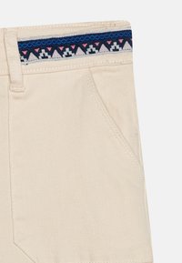 Pantaloni corti beige con una vita a righe blu navy che presenta dettagli rosa e grigio chiaro, e una tasca laterale. Tessuto liscio e resistente.