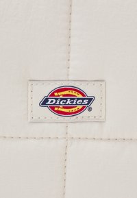Beżowy materiał o pikowanej fakturze z prostokątną metką z logo "Dickies" w kolorach niebieskim, czerwonym i żółtym.