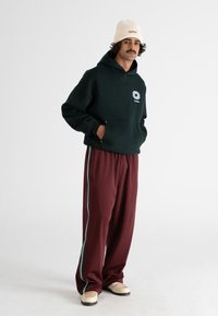 Felpa verde con cappuccio e logo, pantaloni larghi bordeaux con righe turchesi e scarpe beige con tomaia intrecciata.
