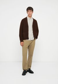 Brown knitted cardigan with buttons and pockets, worn over a light striped shirt, paired with beige trousers and black shoes.
