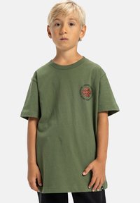 Zelené bavlněné tričko s krátkým rukávem, volnější střih a kulatý výstřih. Na levé straně hrudi se nachází kulatý logo v oranžové a černé barvě.