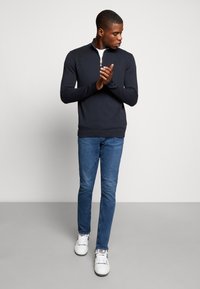 Pulover cu fermoar de marina, dintr-un material moale, purtat împreună cu blugi slim-fit albaștri și adidași albi. Include un guler înalt și tiv cu dungi.