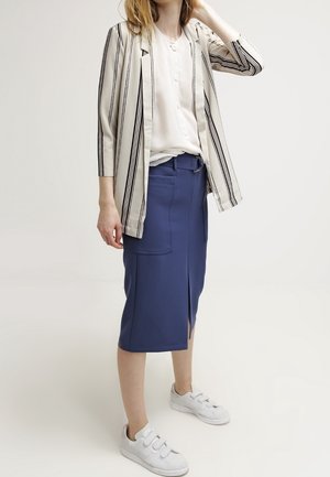 Gestreepte blazer in crème en zwart, witte overhemd, blauwe midi-rok met een vooropening, en witte sneakers.