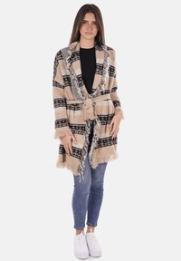 Cardigan lungo beige a quadri con motivi neri, dettagli con frange, apertura frontale e una cintura coordinata. Abbinato a jeans blu e sneakers bianche.