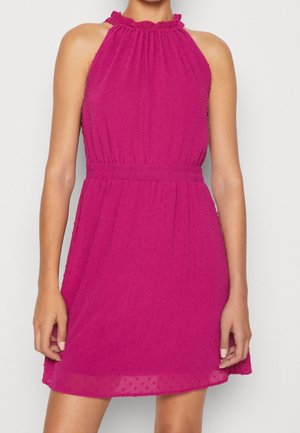 Mujer que lleva un vestido sin mangas de color rosa brillante con un cuello alto con volantes y tela texturizada con puntos, cintura ceñida con detalles fruncidos.