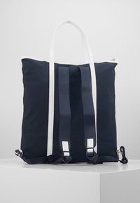 Marinblå tote-väska med vita accenter, utrustad med två axelremmar, dragkedjestängning och en mjuk yta. Metallbeslag synliga.