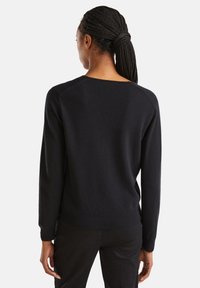 Kobieta z warkoczami związanymi z tyłu, ubrana w zwykły czarny sweter z długim rękawem i czarne spodnie, odwrócona tyłem na białym tle.